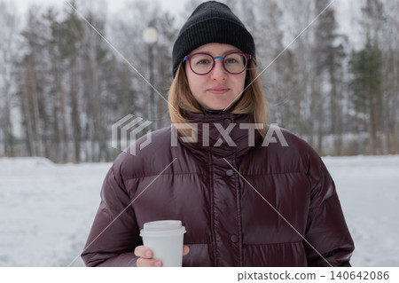 Woman in puffer jacket holding coffee, park setting with snowcovered trees, calm expression with round glasses and knit beanie, warm breath in crisp air, hands wrapped around cup, serene winter pause Woman in puffer jacket holding coffee, park setting with snowcovered trees, calm expression with round glasses and knit beanie, warm breath in crisp air, hands wrapped around cup, serene winter pause 140642086