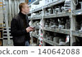 Young electrician browsing electrical cables on smartphone in hardware store aisle surrounded by shelves of goods. A man in a hardware store, choosing electrical wires. 140642088