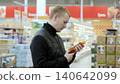 Man carefully reads tomato sauce label in busy supermarket, comparing products with focused expression. Background of blurry shelves highlights mindful shopping experience 140642099