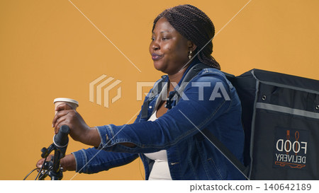 Delivery by active woman riding bicycle around neighborhood while holding coffee in her hand, with courier consumes healthy drink refreshment before delivering food. 140642189