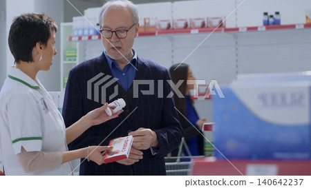 Trained pharmacy assistant helping elderly man with medical counsel, with elderly customer in pharmacy wishing to purchase heartbeat regulating antibiotics, asking expert for guidance. Trained pharmacy assistant helping elderly man with medical counsel, with elderly customer in pharmacy wishing to purchase heartbeat regulating antibiotics, asking expert for guidance. 140642237