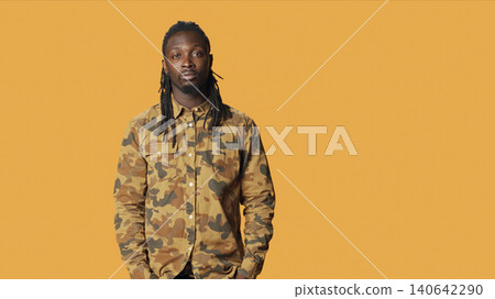 Man with camo shirt posing in studio, smiling and feeling confident over orange background, with young male model feeling joyful and excited on camera, expressing positivity. Man with camo shirt posing in studio, smiling and feeling confident over orange background, with young male model feeling joyful and excited on camera, expressing positivity. 140642290