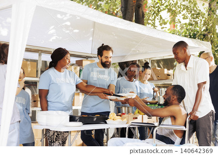 Free meals and fruits are given to a wheelchair-bound man in need, with smiling volunteers of different ethnicities aiding the handicapped poor and homeless people at outdoor food bank. Free meals and fruits are given to a wheelchair-bound man in need, with smiling volunteers of different ethnicities aiding the handicapped poor and homeless people at outdoor food bank. 140642369