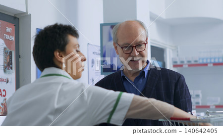 Elderly man arriving at pharmacy checkout counter after finding needed medical items, buying disease cure pills with credit card, being helped by pharmacist with shopping bag. Elderly man arriving at pharmacy checkout counter after finding needed medical items, buying disease cure pills with credit card, being helped by pharmacist with shopping bag. 140642405