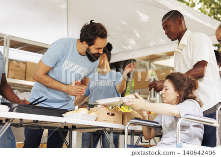 Friendly male volunteer assists and serves free food to the poor needy woman in wheelchair. Friendly male volunteer assists and serves free food to the poor needy woman in wheelchair. 140642406