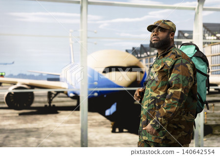 Man in camouflage clothing walking in airport terminal windows with large backpack, next to aircrafts visible behind windows. African american army soldier in airport walking to plane Man in camouflage clothing walking in airport terminal windows with large backpack, next to aircrafts visible behind windows. African american army soldier in airport walking to plane 140642554
