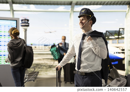 Airline captain in uniform walking through busy airport terminal with suitcase, preparing for flight. Professional pilot embody aviation career, reliable service within cabin crew operations. Airline captain in uniform walking through busy airport terminal with suitcase, preparing for flight. Professional pilot embody aviation career, reliable service within cabin crew operations. 140642557