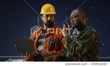 Military man on drilling rig speaks into walkie talkie, enforcing maritime blockade protocol. Soldier on offshore platform talks in handheld radio to receive update on war causing oil crisis, camera A Military man on drilling rig speaks into walkie talkie, enforcing maritime blockade protocol. Soldier on offshore platform talks in handheld radio to receive update on war causing oil crisis, camera A 140642687