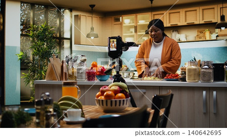 Woman influencer chef recording a food vlog and cutting iceberg salad, kitchen studio with natural light. African American content creator explains the cooking process and preparation. 140642695