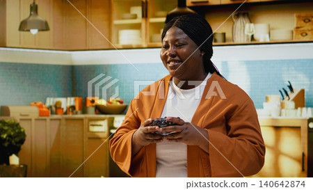 Food vlogger, smelling blueberries, bowl in hand, benefits of the fruits, recording a video at the kitchen counter for online content. Black influencer filming vlog with meal prep advice. Food vlogger, smelling blueberries, bowl in hand, benefits of the fruits, recording a video at the kitchen counter for online content. Black influencer filming vlog with meal prep advice. 140642874