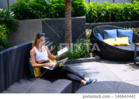 Woman working on laptop computer and reading notebook outdoor lounge, relaxed remote work lifestyle Woman working on laptop computer and reading notebook outdoor lounge, relaxed remote work lifestyle 140643442