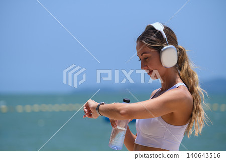 Sporty woman checking her smartwatch while holding a water bottle on the beach Sporty woman checking her smartwatch while holding a water bottle on the beach 140643516