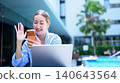 Woman video calling on smartphone by pool while working on laptop outdoors 140643564