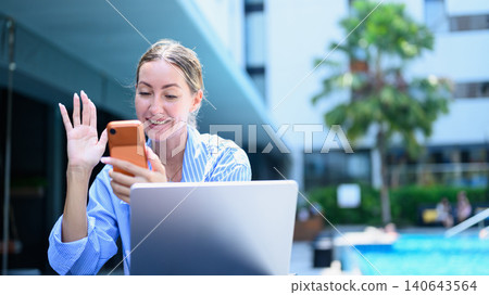 Woman video calling on smartphone by pool while working on laptop outdoors Woman video calling on smartphone by pool while working on laptop outdoors 140643564