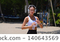 Beautiful sporty woman with headphones holding water bottle on the beach 140643569