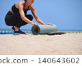 Closeup woman preparing yoga mat on sandy beach, outdoor workout and wellness lifestyle with calm ocean horizon 140643602
