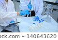 Scientists in White Lab Coats and Blue Gloves Conducting Laboratory Experiment with Test Tubes and Vials 140643620