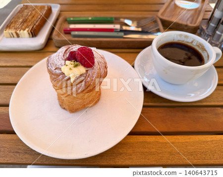 Fresh kraffin with cream topping, raspberry sits beside black coffee on wooden cafe table, honey cake in background, breakfast, bakery atmosphere. Cafe menu, food, pastry, coffee culture, hospitality Fresh kraffin with cream topping, raspberry sits beside black coffee on wooden cafe table, honey cake in background, breakfast, bakery atmosphere. Cafe menu, food, pastry, coffee culture, hospitality 140643715