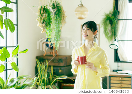 A young Japanese woman drinking a beverage by the window in her living room, surrounded by houseplants. A young Japanese woman drinking a beverage by the window in her living room, surrounded by houseplants. 140643953