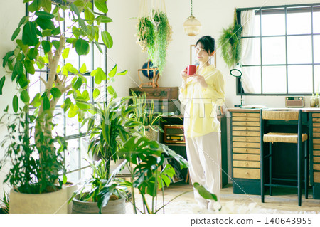 一位年輕的日本女子在客廳窗邊喝飲料,周圍擺放著盆栽。 一位年輕的日本女子在客廳窗邊喝飲料,周圍擺放著盆栽。 140643955