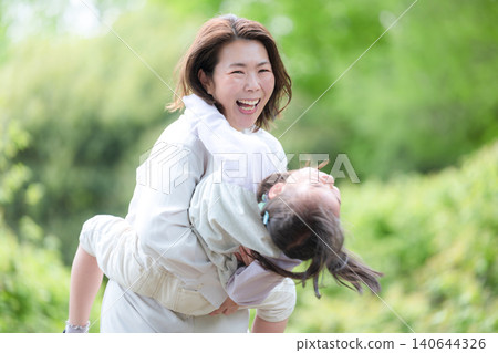 A little girl, smiling and enjoying physical contact with her mother, is held in a park surrounded by fresh greenery. A picture of parent-child love and happy family time. A little girl, smiling and enjoying physical contact with her mother, is held in a park surrounded by fresh greenery. A picture of parent-child love and happy family time. 140644326