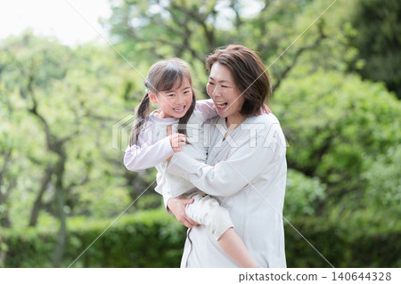 A little girl, smiling and enjoying physical contact with her mother, is held in a park surrounded by fresh greenery. A picture of parent-child love and happy family time. A little girl, smiling and enjoying physical contact with her mother, is held in a park surrounded by fresh greenery. A picture of parent-child love and happy family time. 140644328