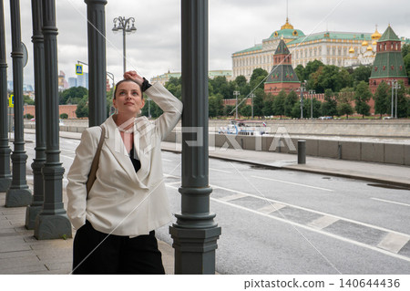 Woman stands on Sofiyskaya Embankment in Moscow with Kremlin across river, framed by colonnade and overcast sky in contemporary urban setting. Travel destination, city architecture, cultural heritage 140644436
