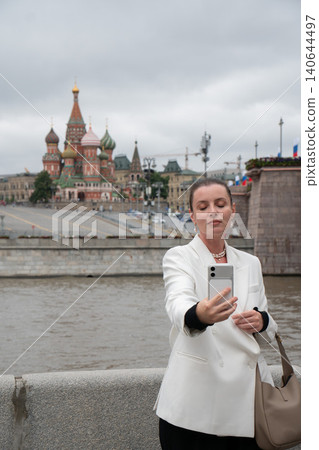 Woman takes smartphone selfie near Moskva River with Saint Basils Cathedral in background, travel experience, iconic architecture, contemporary city lifestyle. Social media, destination discovery Woman takes smartphone selfie near Moskva River with Saint Basils Cathedral in background, travel experience, iconic architecture, contemporary city lifestyle. Social media, destination discovery 140644497