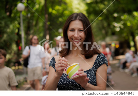 A cheerful woman stands in a sunlit park, holding a colorful striped cup and smiling as pedestrians pass by in the blurred background amid green trees A cheerful woman stands in a sunlit park, holding a colorful striped cup and smiling as pedestrians pass by in the blurred background amid green trees 140644945