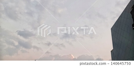 The afternoon sky is filled with thin gray and faded orange clouds. In the right corner is the side of a modern gray building with a checkered pattern. A calm atmosphere, soft light approaching evenin The afternoon sky is filled with thin gray and faded orange clouds. In the right corner is the side of a modern gray building with a checkered pattern. A calm atmosphere, soft light approaching evenin 140645750