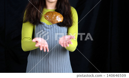 Young woman baking bread and tossing dough in the air, captured mid-motion against a dark background in a kitchen Young woman baking bread and tossing dough in the air, captured mid-motion against a dark background in a kitchen 140646509