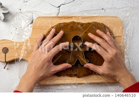 Hands pressing a gingerbread man cookie cutter into rolled dough on a wooden board, preparing homemade Christmas holiday cookies Hands pressing a gingerbread man cookie cutter into rolled dough on a wooden board, preparing homemade Christmas holiday cookies 140646510