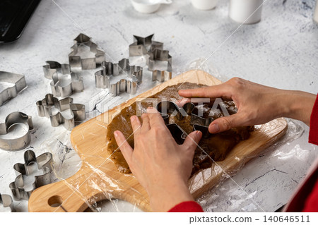 Hands using a gingerbread man cookie cutter on rolled dough surrounded by festive molds, preparing homemade Christmas cookies in the kitchen Hands using a gingerbread man cookie cutter on rolled dough surrounded by festive molds, preparing homemade Christmas cookies in the kitchen 140646511