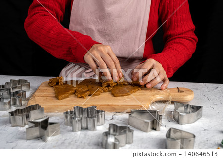 Person in a red sweater and apron separating gingerbread cookie shapes from dough on a wooden board during holiday baking Person in a red sweater and apron separating gingerbread cookie shapes from dough on a wooden board during holiday baking 140646513