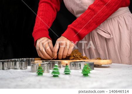Person in a red sweater and apron shaping gingerbread dough on a wooden board surrounded by cookie cutters and festive decorations Person in a red sweater and apron shaping gingerbread dough on a wooden board surrounded by cookie cutters and festive decorations 140646514