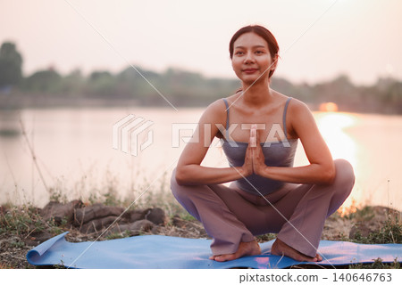 Woman holding deep squat yoga pose with prayer hands on a blue mat beside a lake at sunset, highlighting outdoor mindfulness, hip mobility, balance, grounding, flexibility, and healthy lifestyle. Woman holding deep squat yoga pose with prayer hands on a blue mat beside a lake at sunset, highlighting outdoor mindfulness, hip mobility, balance, grounding, flexibility, and healthy lifestyle. 140646763