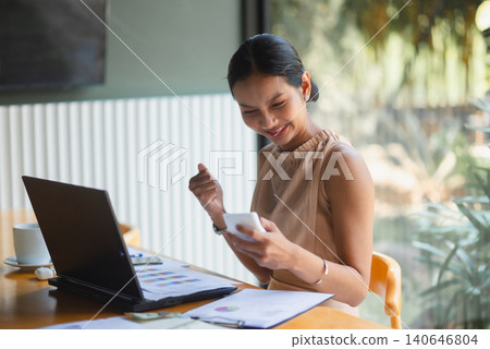 Smiling businesswoman using a smartphone beside laptop, charts, cash, and paperwork at an office desk, representing online finance, budgeting success, accounting review, digital work, Smiling businesswoman using a smartphone beside laptop, charts, cash, and paperwork at an office desk, representing online finance, budgeting success, accounting review, digital work, 140646804