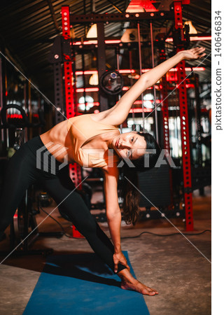 Woman holding triangle yoga pose on a blue mat in a dark gym, emphasizing flexibility, balance, side stretch, core strength, body alignment, mindful movement, wellness, and fitness routine. Woman holding triangle yoga pose on a blue mat in a dark gym, emphasizing flexibility, balance, side stretch, core strength, body alignment, mindful movement, wellness, and fitness routine. 140646834