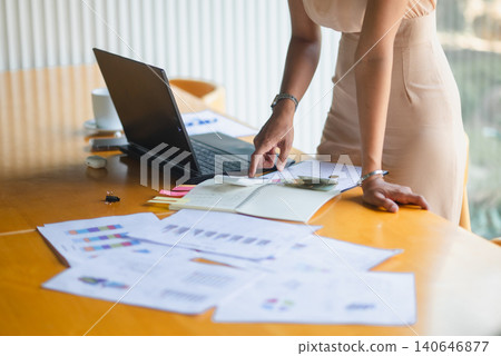 Businesswoman standing at a desk using a calculator with laptop, notebook, cash, and financial reports, representing accounting work, budget review, expense planning, and office productivity. Businesswoman standing at a desk using a calculator with laptop, notebook, cash, and financial reports, representing accounting work, budget review, expense planning, and office productivity. 140646877