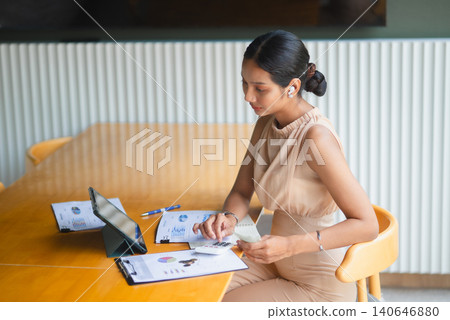 Businesswoman wearing earbuds uses a calculator beside tablet, notebook, cash, and financial charts, representing remote work, online finance, budgeting, accounting, and data review. Businesswoman wearing earbuds uses a calculator beside tablet, notebook, cash, and financial charts, representing remote work, online finance, budgeting, accounting, and data review. 140646880