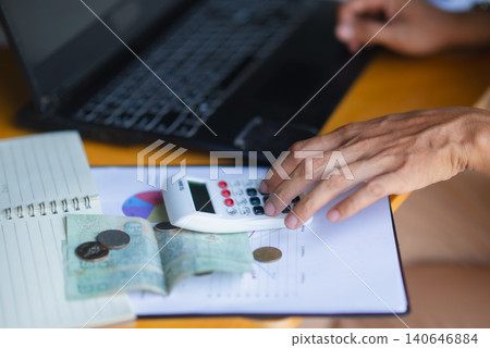 Close-up of hands using a calculator beside laptop, cash, coins, and financial charts on a desk, showing budget calculation, accounting, payment planning, savings, and expense review. Close-up of hands using a calculator beside laptop, cash, coins, and financial charts on a desk, showing budget calculation, accounting, payment planning, savings, and expense review. 140646884