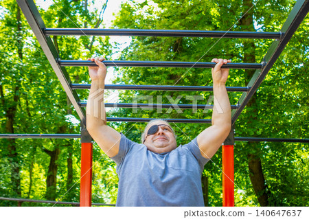 Middle-aged man with eyepatch doing pull-up on monkey bars in outdoor park fitness area Middle-aged man with eyepatch doing pull-up on monkey bars in outdoor park fitness area 140647637