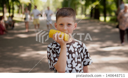 A young boy wearing a patterned shirt stands in a bright park, eating corn on the cob 140648711