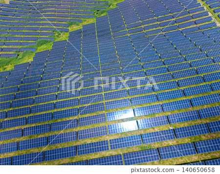 Sunlit reflective panels form geometric pattern over lush landscape. Aerial view captures shimmering solar arrays creating organized blue patches on vibrant grass Sunlit reflective panels form geometric pattern over lush landscape. Aerial view captures shimmering solar arrays creating organized blue patches on vibrant grass 140650658