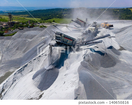 Heavy machinery works in a granite quarry with piles of stone and dust in the air during bright daylight hours near a mountainous area 140650816