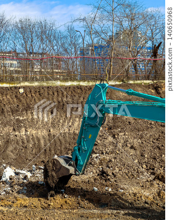 Construction machine digs into dirt at a construction site near a city with buildings in the background on a clear day Construction machine digs into dirt at a construction site near a city with buildings in the background on a clear day 140650968