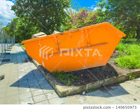Bright orange dumpster stands on the side of a sidewalk with greenery and trees in the background under a clear sky Bright orange dumpster stands on the side of a sidewalk with greenery and trees in the background under a clear sky 140650973
