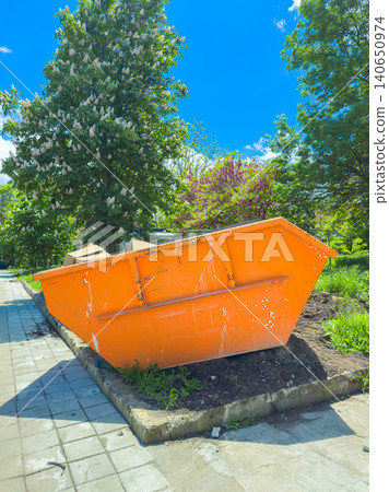 Construction site with an orange skip container on a sunny day in a park with trees and green plants, showing ongoing renovation and maintenance work near a walking path Construction site with an orange skip container on a sunny day in a park with trees and green plants, showing ongoing renovation and maintenance work near a walking path 140650974