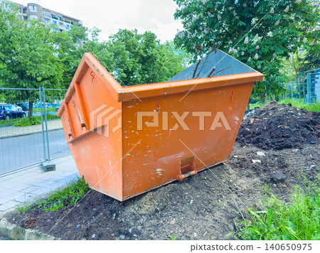 Construction debris bin placed on a grassy area near a street with trees and buildings in the background during daytime hours Construction debris bin placed on a grassy area near a street with trees and buildings in the background during daytime hours 140650975