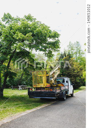 Park maintenance truck beside picnic area with yellow cage and folded crane arm, lush canopy overhead, bright exposure, community stewardship vibe, environmental upkeep 140651012